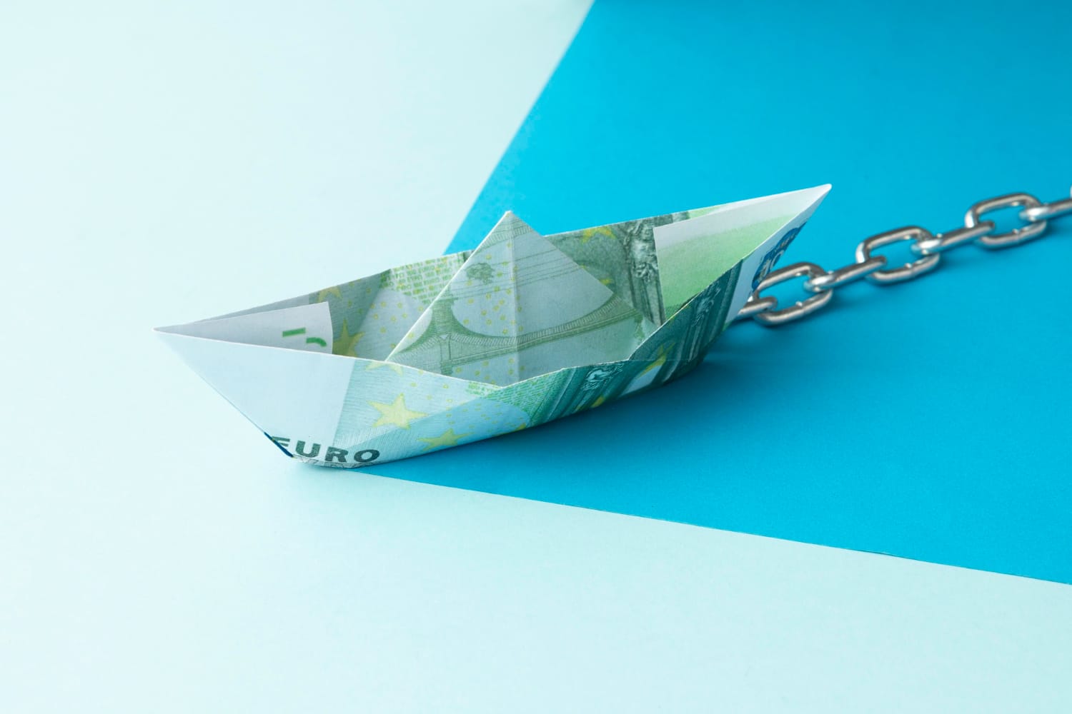 Paper boat made from euro currency chained on blue background symbolizing financial constraints and investment challenges in water infrastructure and pricing